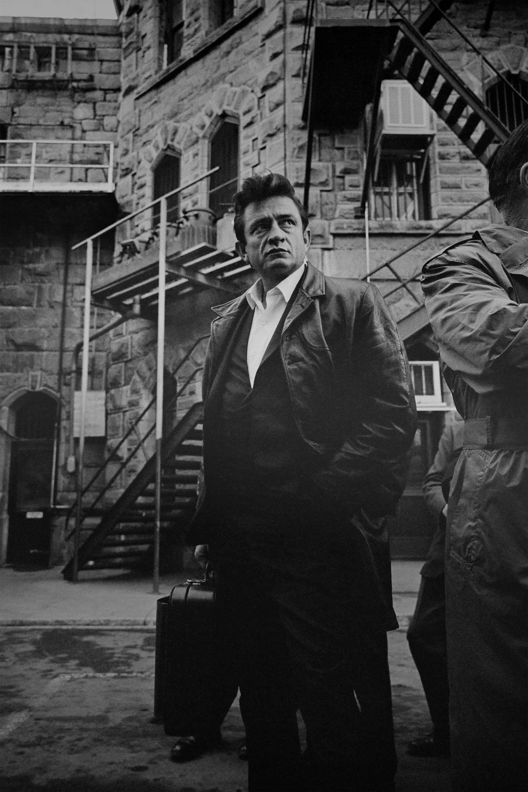 Man in leather jacket stands with suitcase outside stone building with fire escapes, black and white.