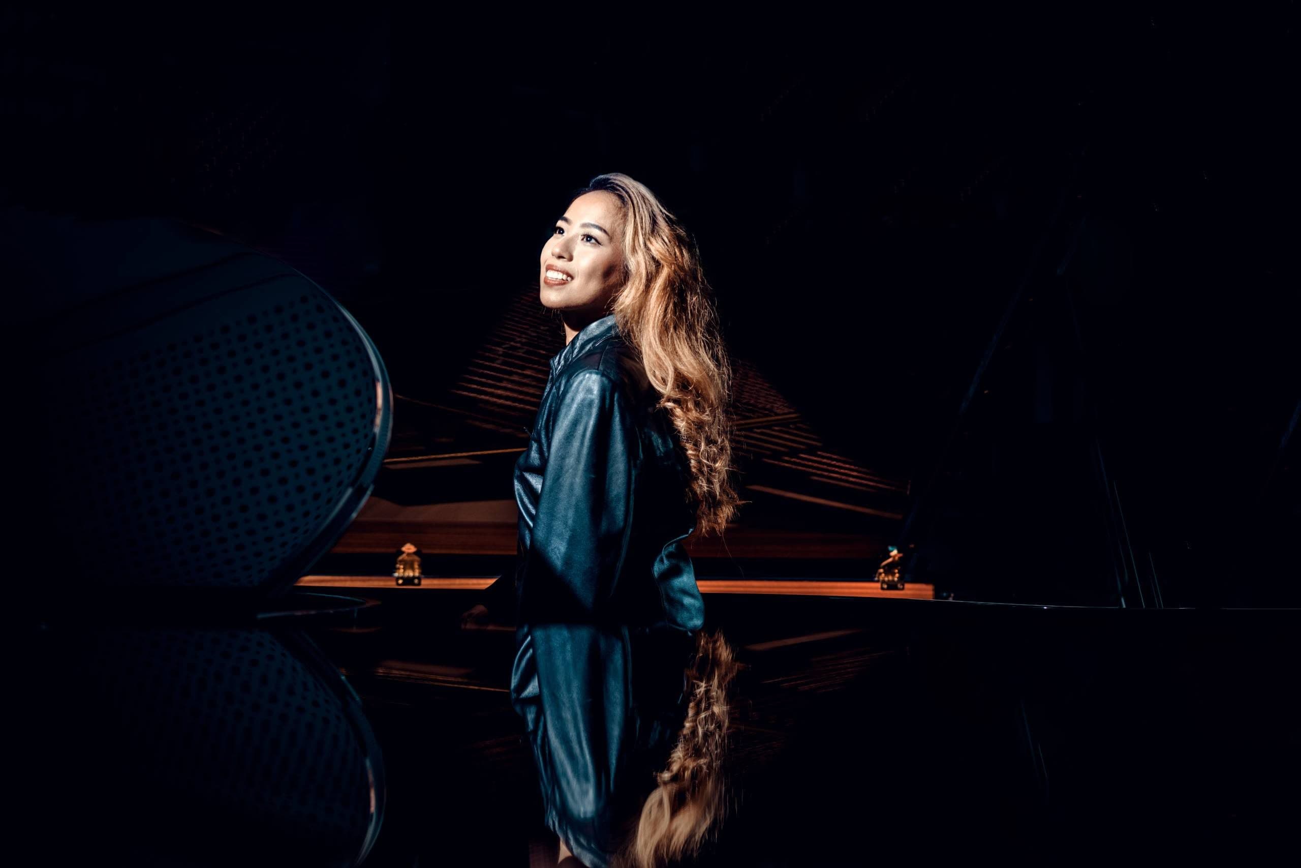 Woman in dark leather jacket standing beside a grand piano in a dimly lit room with reflection.